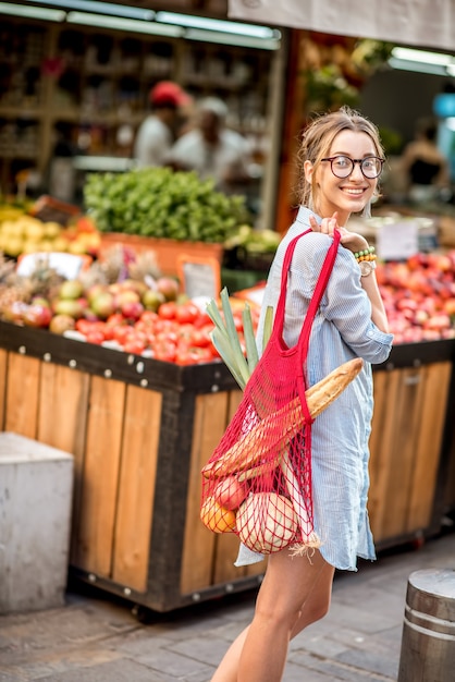 Mujer joven de pie al aire libre con una bolsa de malla llena de verduras frescas frente al mercado de alimentos