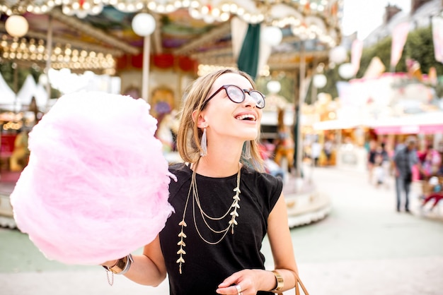 Mujer joven de pie con algodón de azúcar rosa al aire libre frente al carrusel en el parque de atracciones