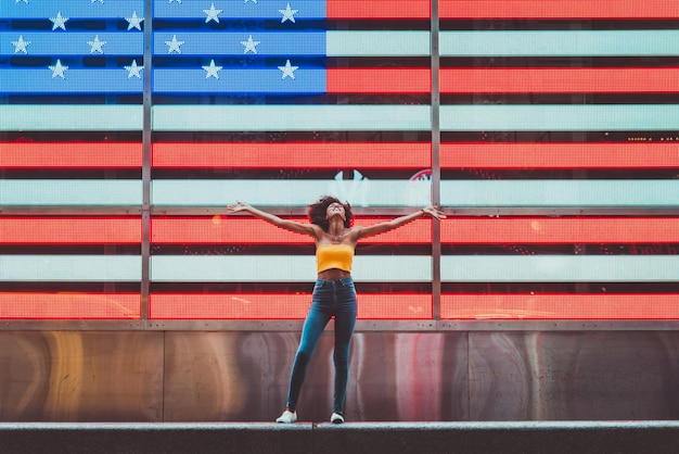Foto mujer joven de pie con los brazos extendidos contra la bandera americana de neón en la ciudad