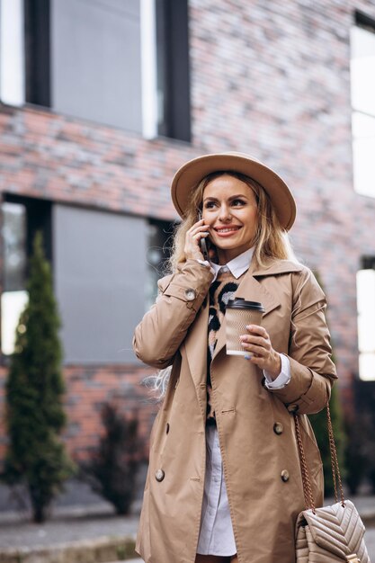 Mujer joven de pie en la calle y tomando café y usando el teléfono