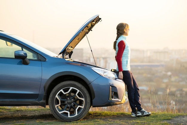 Mujer joven de pie cerca de un coche averiado con un capó abierto que tiene problemas con su vehículo