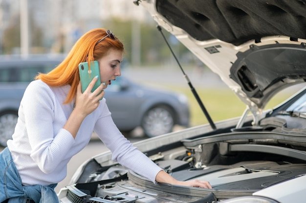 Mujer joven de pie cerca de un coche roto con capucha reventada hablando por su teléfono móvil.