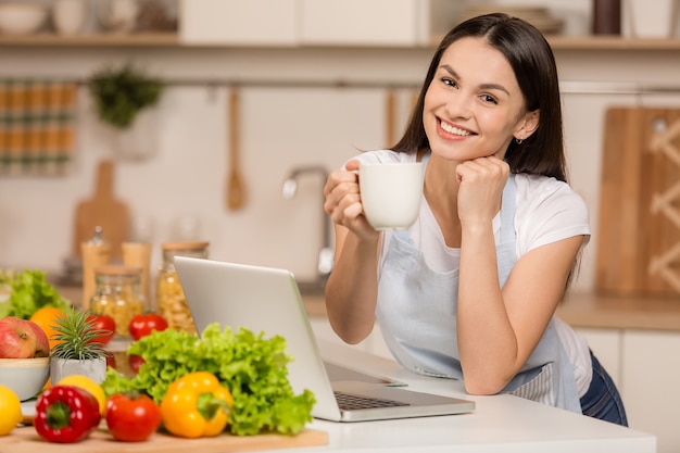 Mujer joven de pie cerca del escritorio en la cocina, sonriendo, con una taza de té