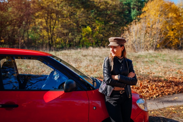 Foto mujer joven de pie en coche en otoño camino. el conductor se detuvo automáticamente en el bosque para disfrutar del paisaje de otoño