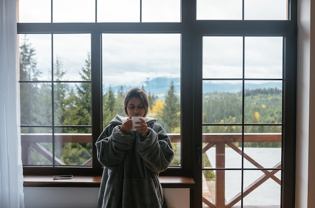 Mujer joven de pie junto a la ventana sostiene una taza