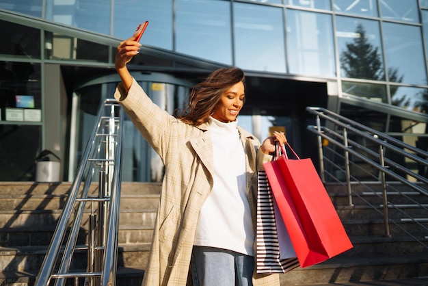 Mujer joven de pie a lo largo de la carretera sosteniendo bolsas de compras usando teléfono móvil Concepto de venta de descuentos