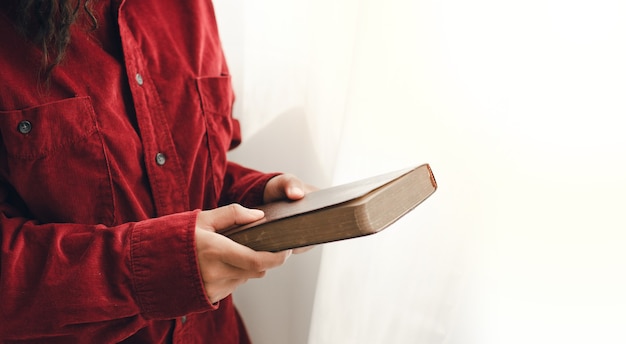 Una mujer joven de pie y orando en la Biblia, copie el espacio. Al lado de la ventana espejada