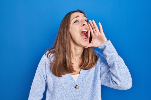 Mujer joven de pie sobre fondo azul gritando y gritando fuerte a un lado con el concepto de comunicación de la mano en la boca