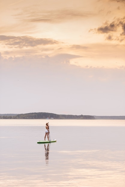 Mujer joven en pie en la tabla de paddle sup con hermosos colores al atardecer o al amanecer
