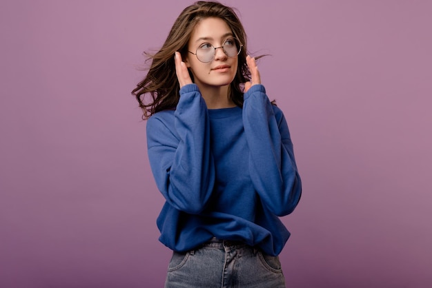 Mujer joven de piel clara con cabello oscuro mira hacia otro lado usa gafas con capucha y jeans sobre fondo púrpura Concepto de confianza en sí mismo