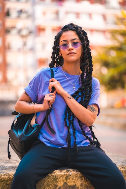 Mujer joven de piel oscura con largas trenzas y gafas moradas sentado en un banco de la ciudad