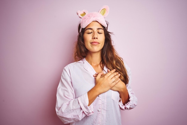 Mujer joven con pijama y antifaz para dormir de pie sobre un fondo rosa aislado sonriendo con las manos en el pecho con los ojos cerrados y un gesto agradecido en la cara Concepto de salud