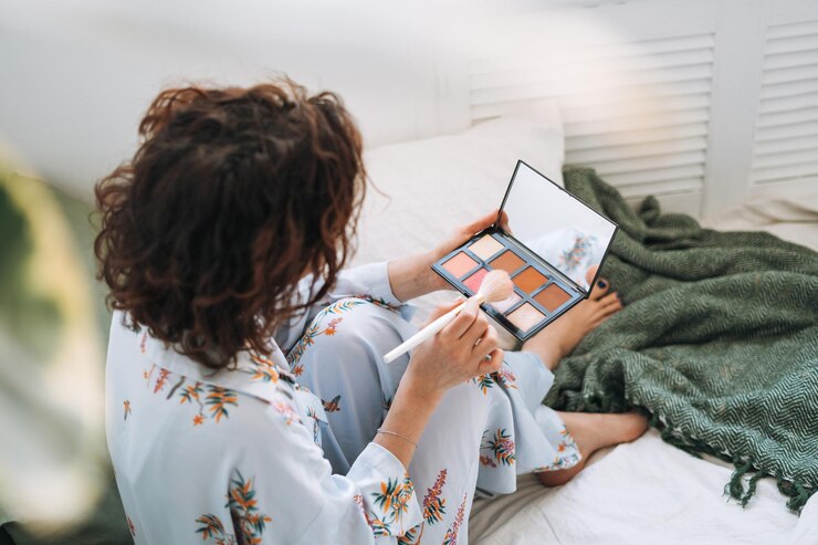 Foto mujer joven en pijama azul aplica maquillaje con paleta de escultura facial sentada en la cama en casa