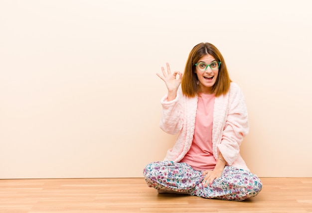 Mujer joven con pijama sentada en casa sintiéndose exitosa y satisfecha, sonriendo con la boca abierta, haciendo un signo bien con la mano