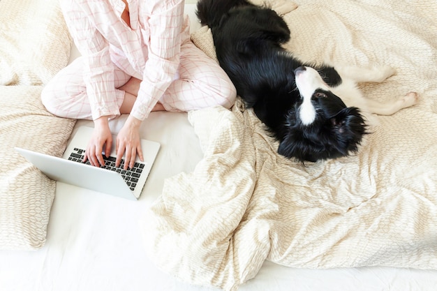 Mujer joven en pijama sentado en la cama con perro trabajando usando la computadora portátil pc en casa