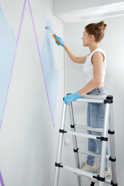 Mujer joven pintando la pared con rodillo de pintura y usando cinta adhesiva mientras está de pie en la escalera