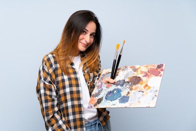 Mujer joven pintor sobre pared azul aislada con expresión feliz