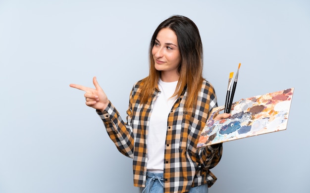 Mujer joven pintor sobre pared azul aislado apuntando hacia un lado para presentar un producto