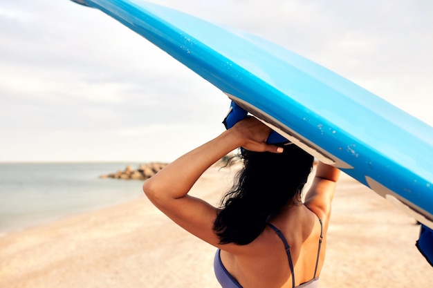 Foto mujer joven en la playa de arena lleva una tabla de surf en la cabeza