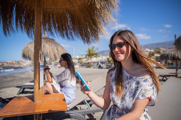 Una mujer joven en la playa usa su teléfono mientras está sentada en la tumbona con su amiga