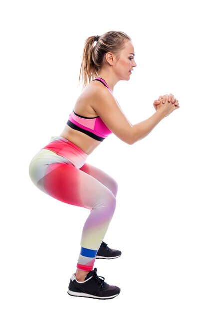 Una mujer joven se pone en cuclillas con una banda elástica en las piernas Mujer de belleza sonriente en ropa deportiva brillante Actividad deportiva y salud Altura completa Aislada sobre fondo blanco Vista lateral Vertical