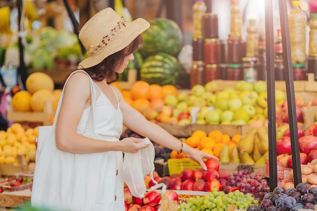 Mujer joven pone frutas y verduras en una bolsa de productos de algodón en el mercado de alimentos. Bolsa ecológica reutilizable para compras. Concepto de desperdicio cero.