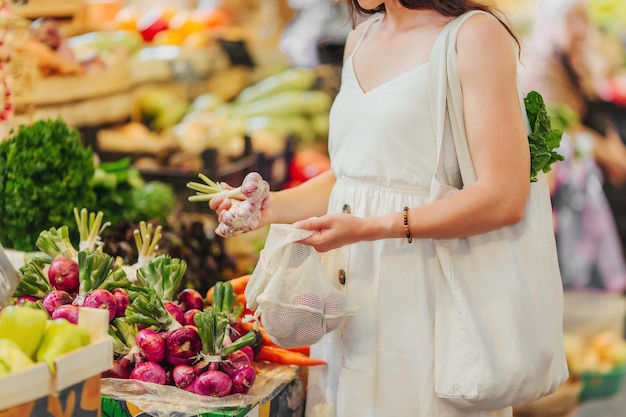 Mujer joven pone frutas y verduras en una bolsa de productos de algodón en el mercado de alimentos. Bolsa ecológica reutilizable para compras. Concepto de desperdicio cero.