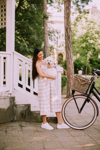 Mujer joven poniendo perro bichon frise blanco en la cesta de la bicicleta eléctrica