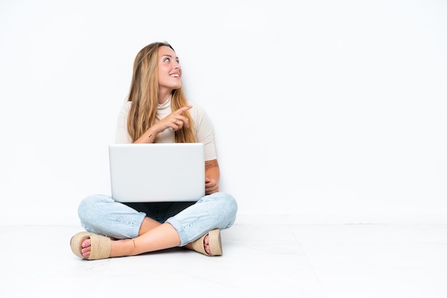 Foto mujer joven con portátil sentada en el suelo aislada de fondo blanco señalando con el dedo al lado y presentando un producto