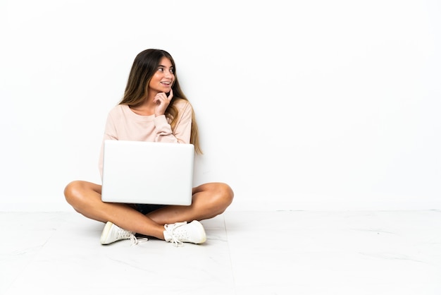 Foto mujer joven con un portátil sentado en el suelo aislado en la pared blanca pensando en una idea mientras mira hacia arriba