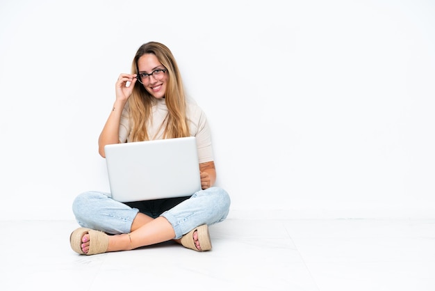 Foto mujer joven con portátil sentado en el suelo aislado sobre fondo blanco con gafas y feliz