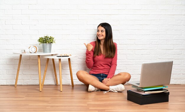 Foto mujer joven con un portátil sentado en el suelo en el interior apuntando hacia un lado para presentar un producto