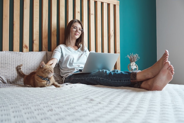 Mujer joven con portátil para trabajar, acostado en la cama con un gato en el dormitorio. Horarios flexibles y trabajo a distancia.