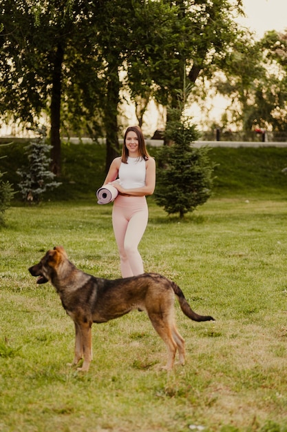 Mujer joven posa en el fondo del parque Morena atractiva en ropa deportiva está de pie sobre la hierba verde al lado del perro y sosteniendo estera de yoga y pilates en sus manos