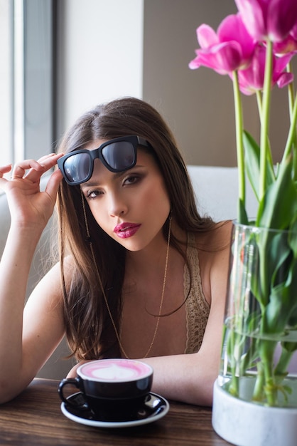 Mujer joven posando en el café Café de la mañana Una taza de capuchino Flores en la mesa Reunión de la mañana