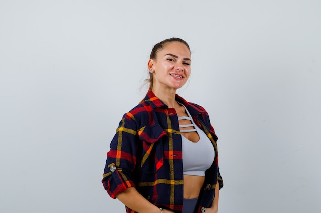 Mujer joven posando mientras está de pie en la parte superior de la cosecha, camisa a cuadros, pantalones y mirando alegre, vista frontal.