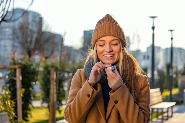 Mujer joven positiva con abrigo cálido sonriendo y mirando hacia otro lado mientras se unta bálsamo labial en los labios durante un paseo por la calle de la ciudad
