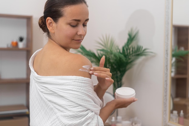 Mujer joven positiva aplicando crema hidratante en el hombro desnudo en el cuidado del cuerpo del baño
