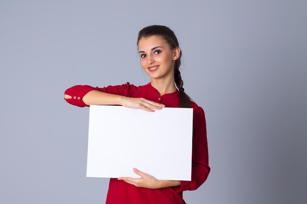 Mujer joven positiva en blusa roja sosteniendo una hoja blanca de papel sobre fondo gris en el estudio