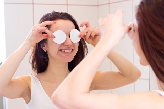 Mujer joven positiva con cabello largo y oscuro en camisa blanca quitando maquillaje con almohadillas de algodón frente al espejo en su baño