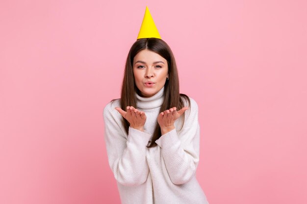 Mujer joven positiva con cabello oscuro en cono de fiesta enviando besos al aire, celebrando cumpleaños, usando suéter blanco de estilo casual. Disparo de estudio interior aislado sobre fondo rosa.