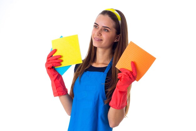 Mujer joven positiva en camiseta azul y delantal con guantes rojos sosteniendo plumeros sobre fondo blanco en el estudio