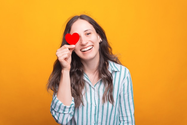 La mujer joven positiva está sonriendo y mirando a la cámara sostiene un corazón de papel cerca de su ojo.