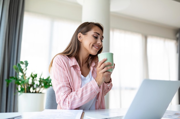 Mujer joven positiva independiente viendo video en blog en netbook y tomando un café en una oficina cómoda Hermosa joven mujer de negocios trabajando en una computadora portátil Freelancer conectándose a internet