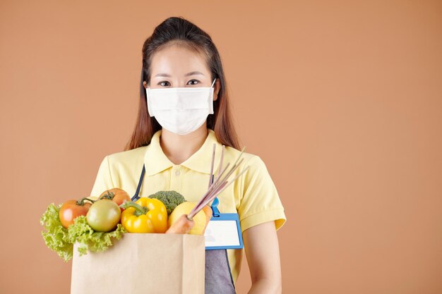 Mujer joven positiva en máscara médica con paquete de papel de comestibles frescos