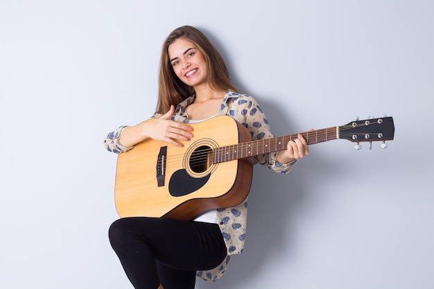Mujer joven positiva con el pelo largo en chaqueta marrón tocando la guitarra en el estudio