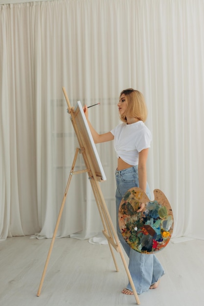 Mujer joven positiva sosteniendo pincel y paleta con pinturas de colores cerca del artista de caballete