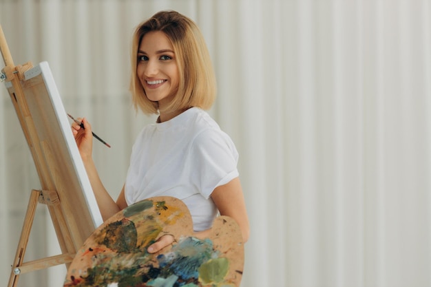 Mujer joven positiva sosteniendo pincel y paleta con pinturas de colores cerca del artista de caballete