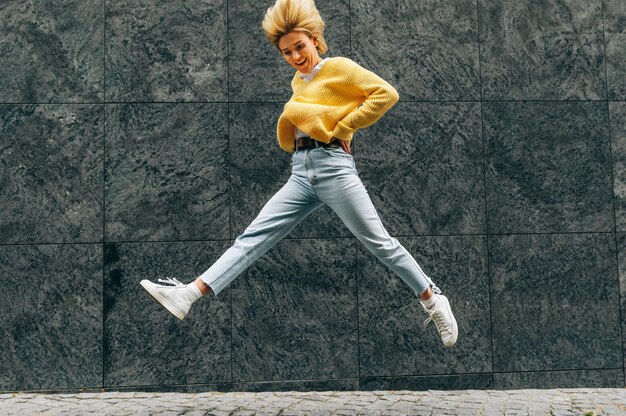 Mujer joven positiva con suéter amarillo azul jeans de cintura alta saltando alto sintiéndose feliz en la calle de la ciudad Mujer rubia estudiante como expresión alegre