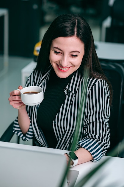 Mujer joven positiva con una taza de café mirando la pantalla del portátil con una sonrisa feliz